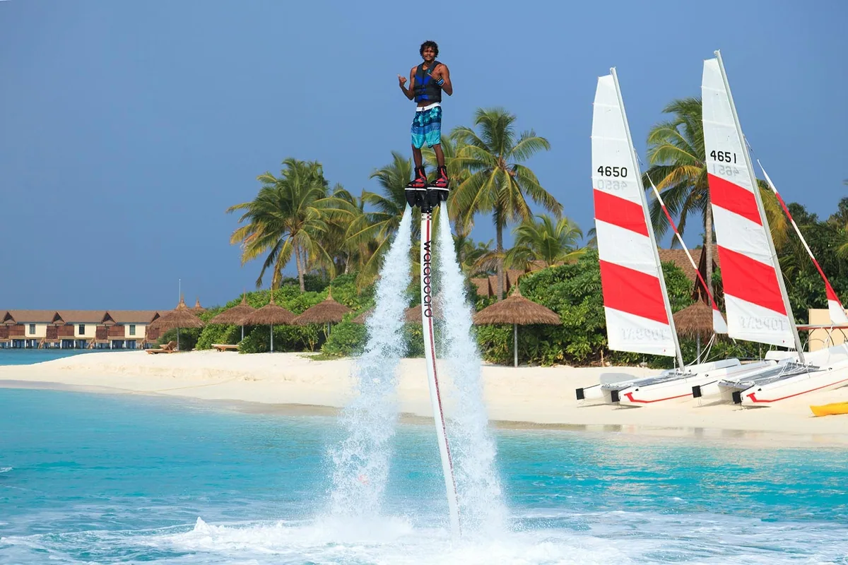 Reethi Faru Resort, activities, Raa Atoll, Maldives, Watersports programme in operation: flyboarder rising over the lagoon under a Flyboard rig, two Hobie Cat catamarans (hull numbers 4650 and 4651) parked on the beach, palm trees framing, the thatched-roof villa row visible in the background.