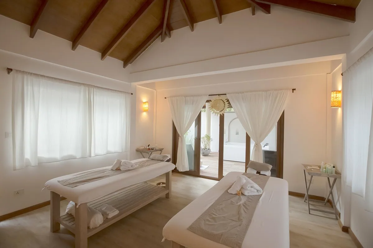 Reethi Faru Resort, spa, Raa Atoll, Maldives, Coconut Spa couples treatment room with bamboo-clad pitched ceiling and exposed wooden rafters, two parallel massage beds with folded white towels, sheer white curtain walls, amber pendant sconces, French doors at the rear opening onto the private outdoor deck with a freestanding tub framed by a sun-disc mirror.