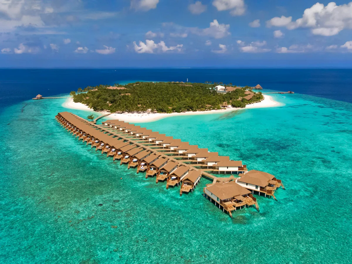 Reethi Faru Resort, setting, Raa Atoll, Maldives, Filaidhoo island aerial alt-angle: palm-covered island with arriving jetty, long overwater villa pier extending forward with thatched-pyramid-roof villas, double-villa terminal pavilion at the pier end, V-shaped arrangement at far-left, clear shallow turquoise lagoon, the reef edge running the full perimeter of the 600-metre island.