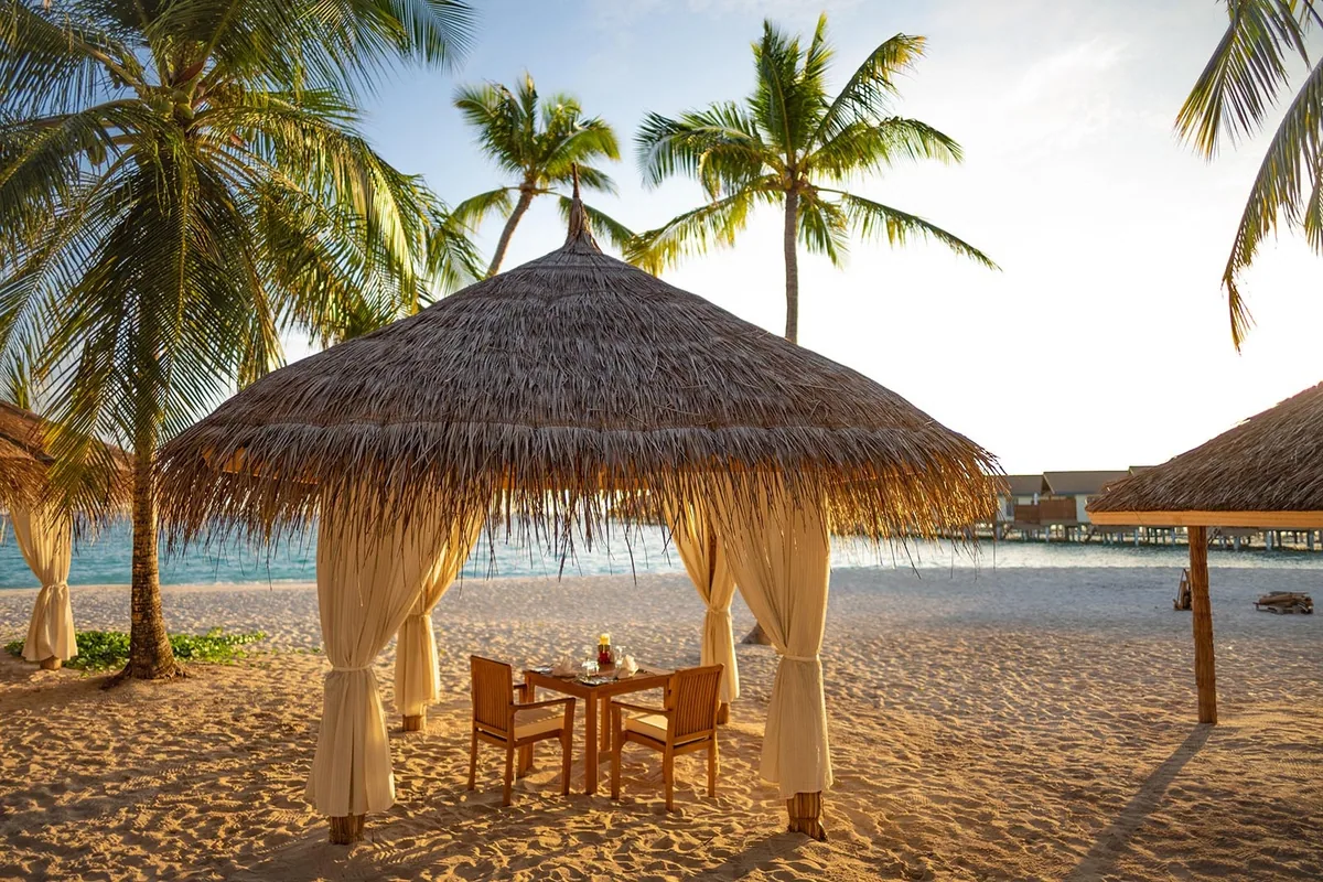 Reethi Faru Resort, dining, Raa Atoll, Maldives, Intimate beach-cabana private dining, single thatched-dome pavilion on the white sand with white draped curtains, table set for two with floral centerpiece, palm fronds on either side and an overwater pavilion in the background in soft evening light.
