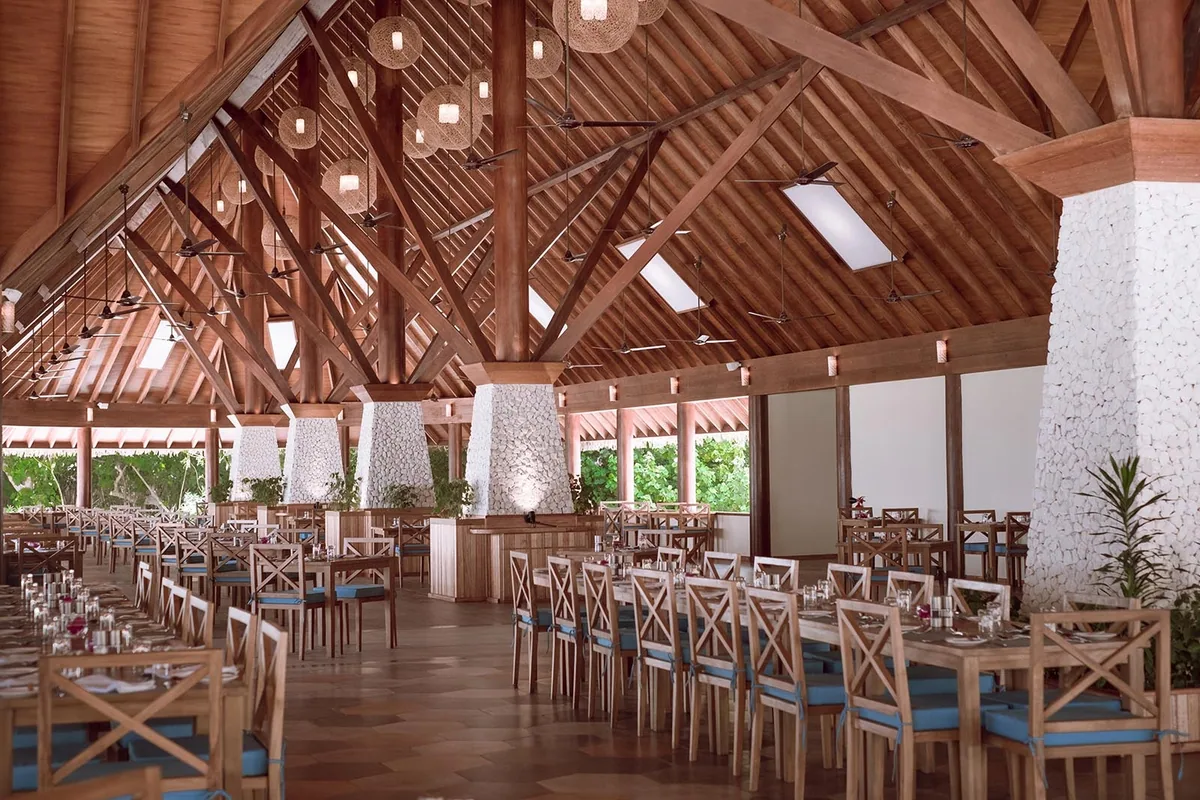 Reethi Faru Resort, dining, Raa Atoll, Maldives, Vakaru main restaurant interior, high-peaked pitched timber-frame roof with exposed rafters and skylights, rope-and-rattan basket pendant lights, coral-pebble-clad columns (Maldivian vernacular signature), long buffet bar in the centre, tables with woven natural-and-blue accent chairs across hardwood flooring.