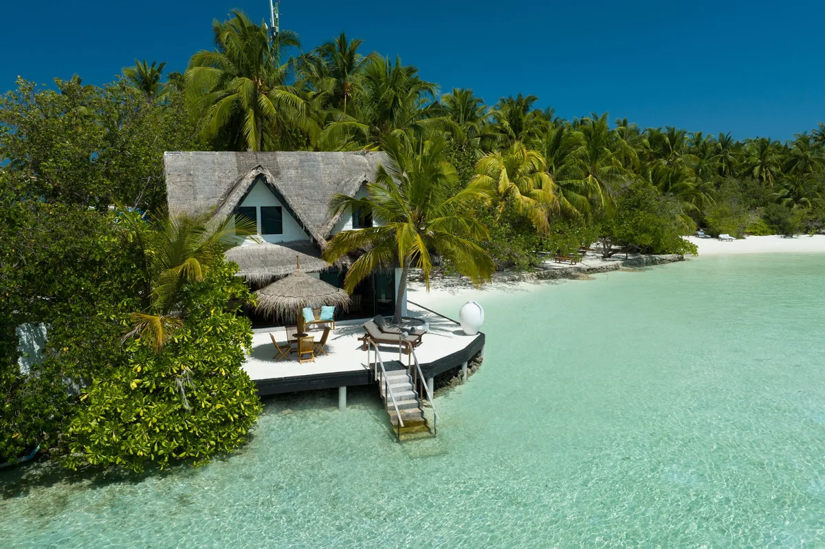Rihiveli Maldives Resort, villa beach, South Malé Atoll, Maldives, Two-Bedroom Family Villa exterior, perched on its own promontory at the island tip with a private wooden deck reaching into the lagoon, four sun-loungers ringed around an A-frame thatched-roof bungalow, ladder down to the turquoise lagoon, dense palm canopy framing the property's signature boutique-honeymoon tier.