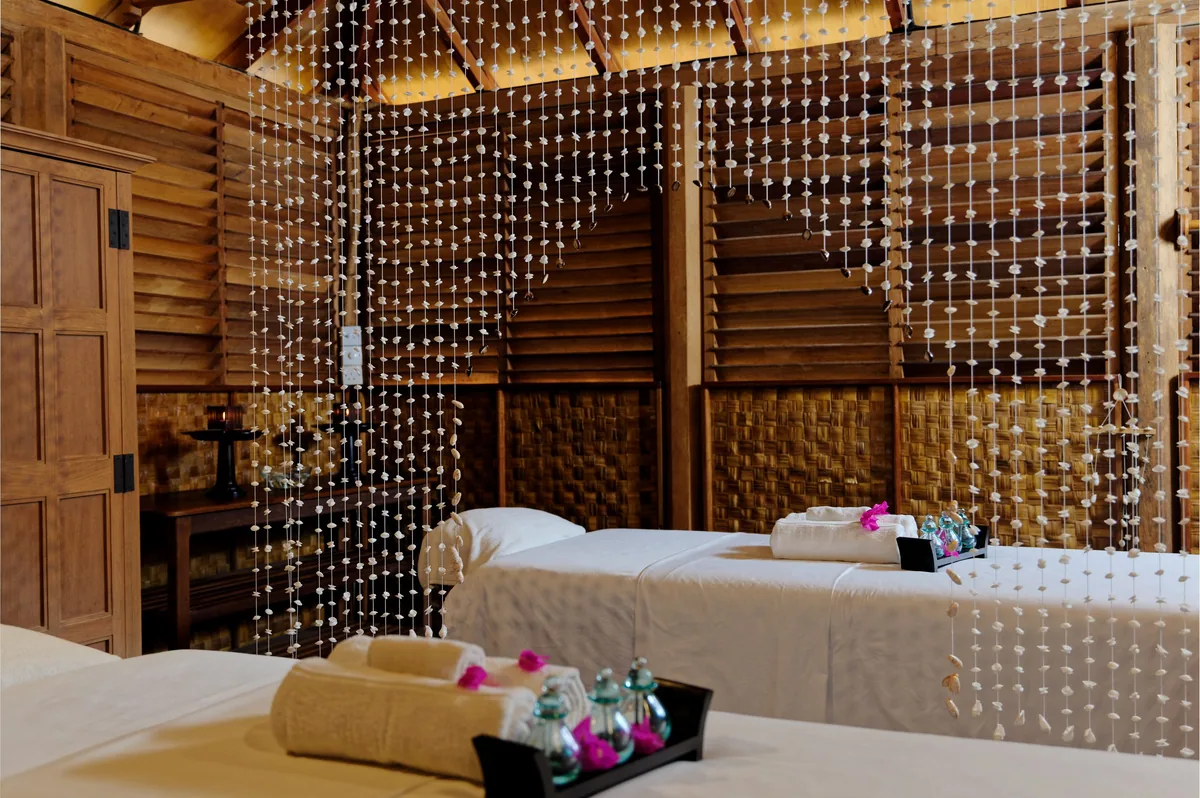 Rihiveli Maldives Resort, spa, South Malé Atoll, Maldives, Spa double-treatment room set up for couples: two parallel massage beds with white linen, dark wood-slat walls, a beaded curtain divider, terra-cotta tile floor, towels and amenity trays with fresh pink frangipani flowers at the bed end, the rustic Maldivian-French aesthetic distinctive to Rihiveli's wellness programme.