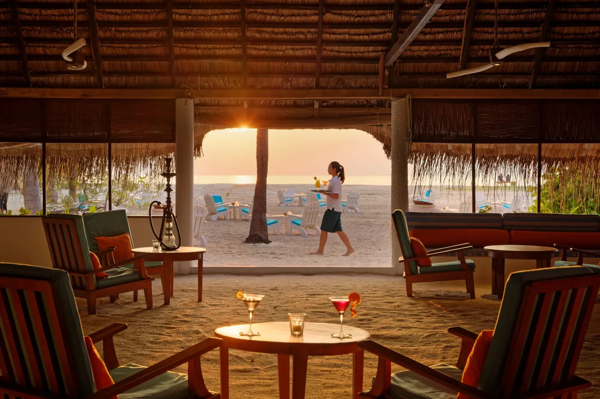 Rihiveli Maldives Resort, dining, South Malé Atoll, Maldives, Sunset Bar interior at the daily French-aperitif cluster: open-air thatched-roof pavilion with sand floor, wooden Adirondack-style lounge chairs, a hookah on the side table, two cocktail glasses on a wooden table in the foreground, server stepping out onto the beach with a tray, white Adirondack chairs arranged on the sand outside, the sun dropping below the horizon.