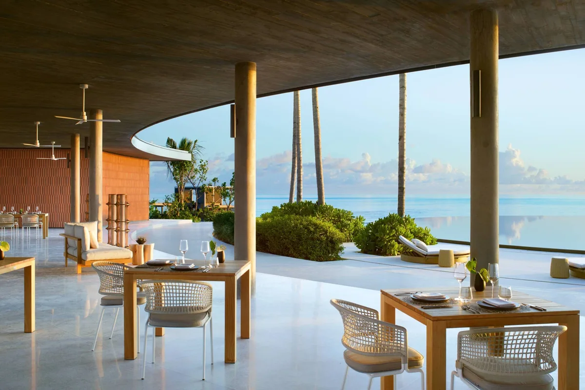 The Ritz-Carlton Maldives, Fari Islands, dining, North Malé Atoll, Maldives, Eau Bar lounge at golden hour: the iconic curved overhanging concrete roof (a Kengo Kuma signature), terracotta-tiled feature wall in the back, woven cane armchairs paired with light wood tables, palm trees framing the reflective infinity-edge surface and the lagoon horizon.