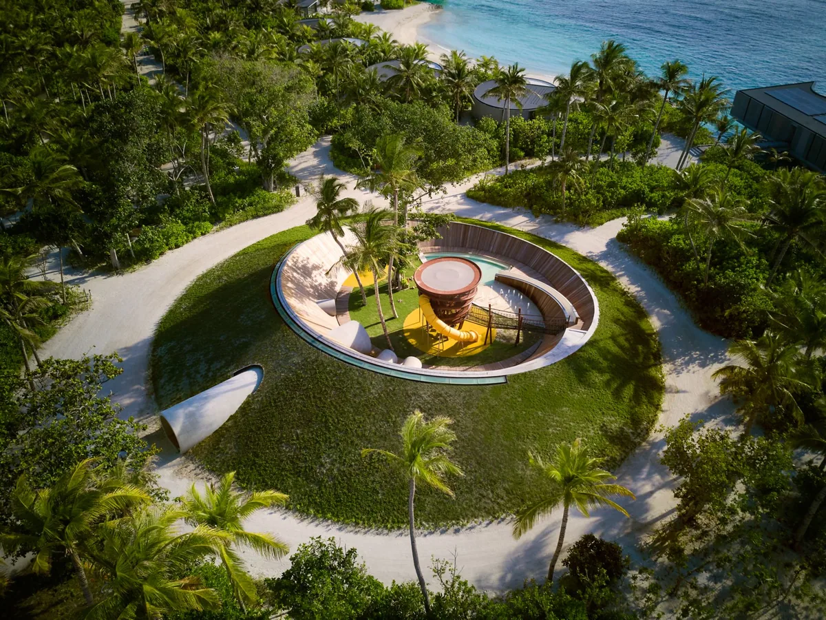 The Ritz-Carlton Maldives, Fari Islands, activity, North Malé Atoll, Maldives, Ritz Kids Club aerial: circular dark-banded outdoor play structure with a bright yellow slide and climbing features at the centre, ringed by sand path and lawn, palm grove enclosure, beach and turquoise lagoon visible at the upper edge. Continues the property's circular geometry.