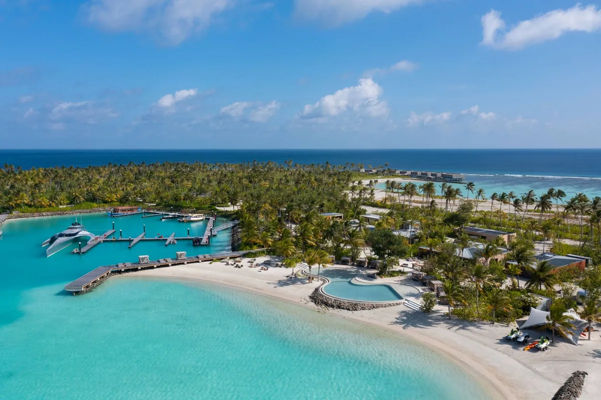 The Ritz-Carlton Maldives, Fari Islands, setting, North Malé Atoll, Maldives, Fari Marina Village aerial: a private yacht docked at the marina jetty, sculpted beachfront with infinity pool nestled in landscaping, jet skis on the sand, palm-covered island stretching to the horizon, the wider Fari Islands commercial precinct anchored by the Ritz-Carlton + Patina cluster.