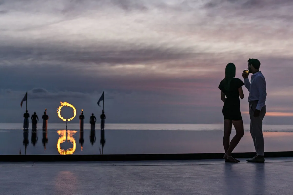 The Ritz-Carlton Maldives, Fari Islands, experience, North Malé Atoll, Maldives, The property's Defining Moment sunset ceremony: a couple silhouetted on a terrace with sundowners, a flaming circular fire-ring (echoing the property's circular geometry) and traditional Maldivian Boduberu drummers standing on the calm lagoon at dusk, soft pastel sky reflecting on the water.