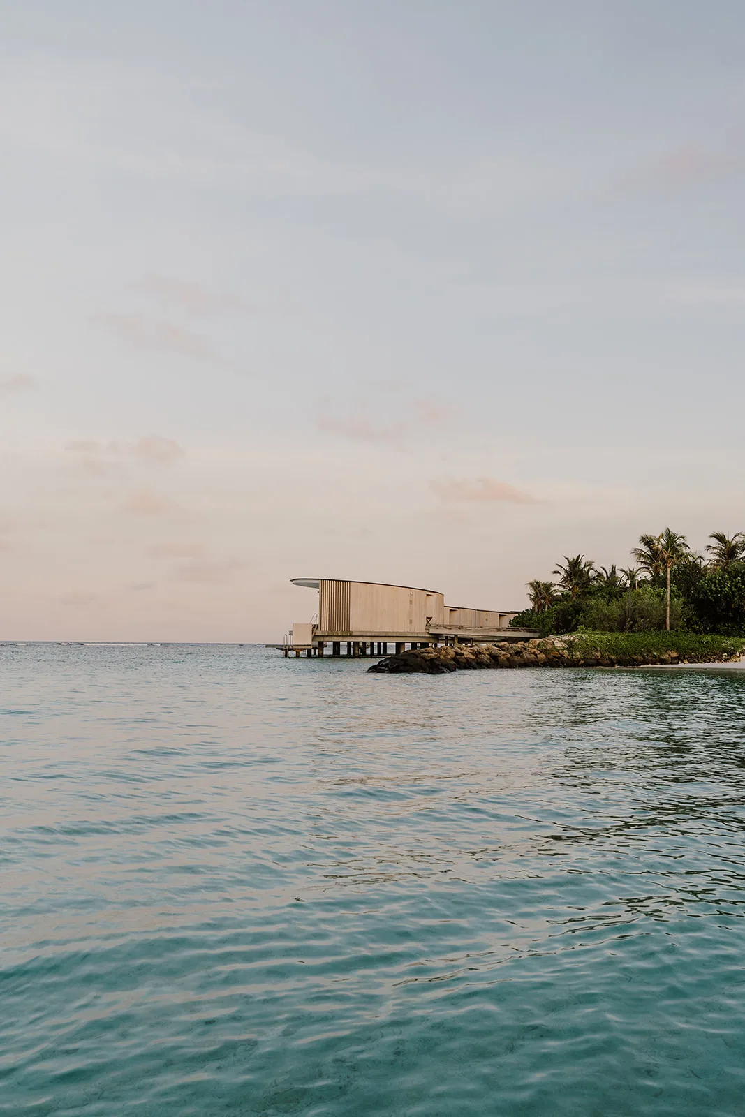 The Ritz-Carlton Maldives, Fari Islands, setting, North Malé Atoll, Maldives, The cylindrical over-water pavilion at the island's edge in dusk pastel light: curved timber-slat exterior on stilts above the lagoon, the soft pink-orange sky reflected on the calm water, palm grove anchoring the shoreline. The cylindrical geometry is Kengo Kuma's signature property vocabulary.