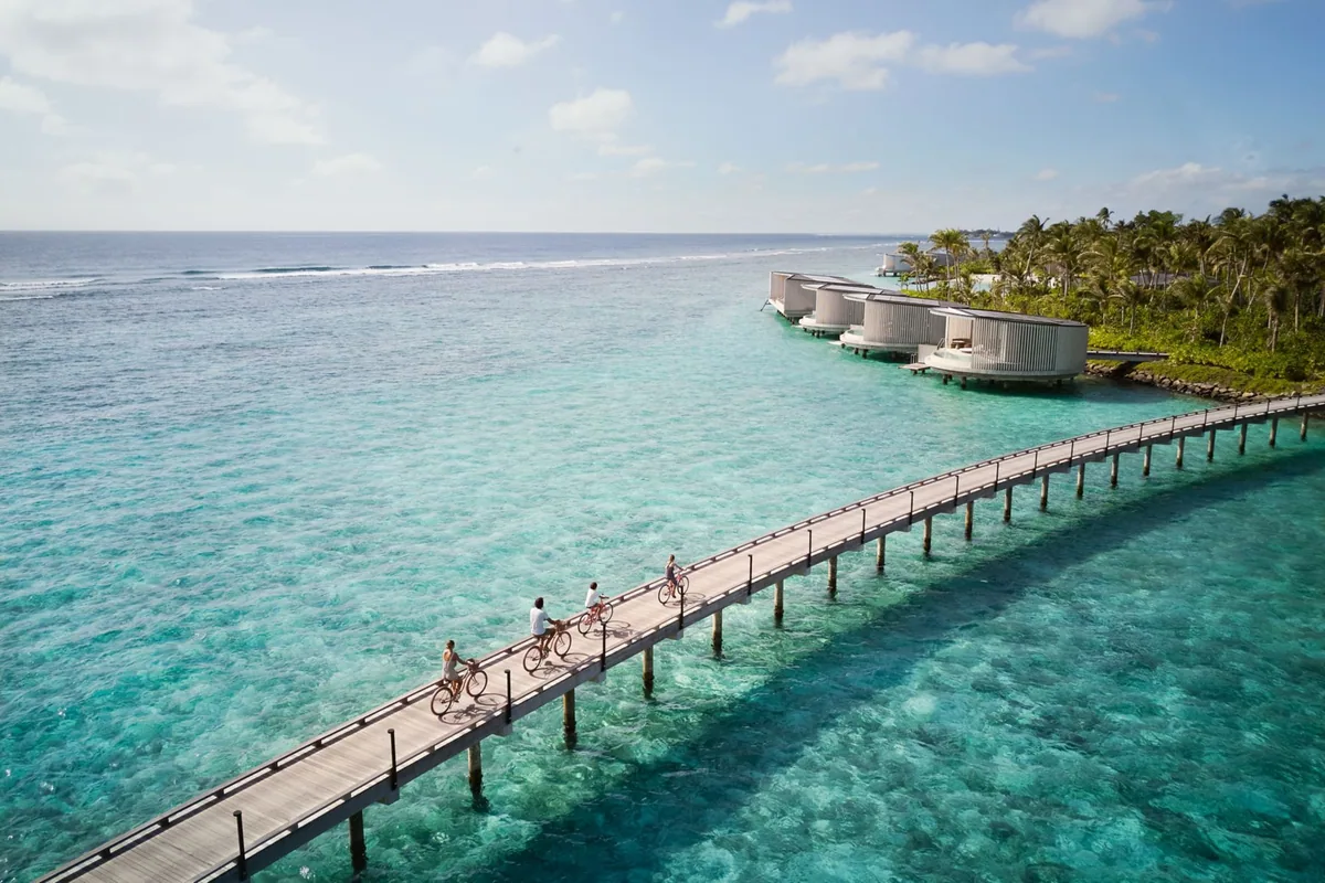 The Ritz-Carlton Maldives, Fari Islands, villa overwater, North Malé Atoll, Maldives, Guests cycling the over-water boardwalk past the property's signature Kengo Kuma cylindrical over-water villa cluster: vertical timber-slat exteriors, palm-fringed beach island in the background, reef-break visible in the distance, North Malé lagoon turquoise underfoot.