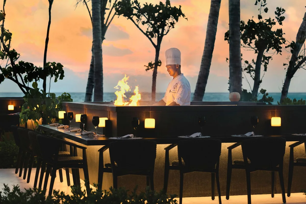 The Ritz-Carlton Maldives, Fari Islands, dining, North Malé Atoll, Maldives, Iwau, the property's Japanese over-water restaurant: open-air teppanyaki counter at sunset with live-flame preparation, chef in traditional whites working the centre grill, paired wine glasses set across the counter, palm-fringed ocean horizon behind, golden-orange dusk sky.