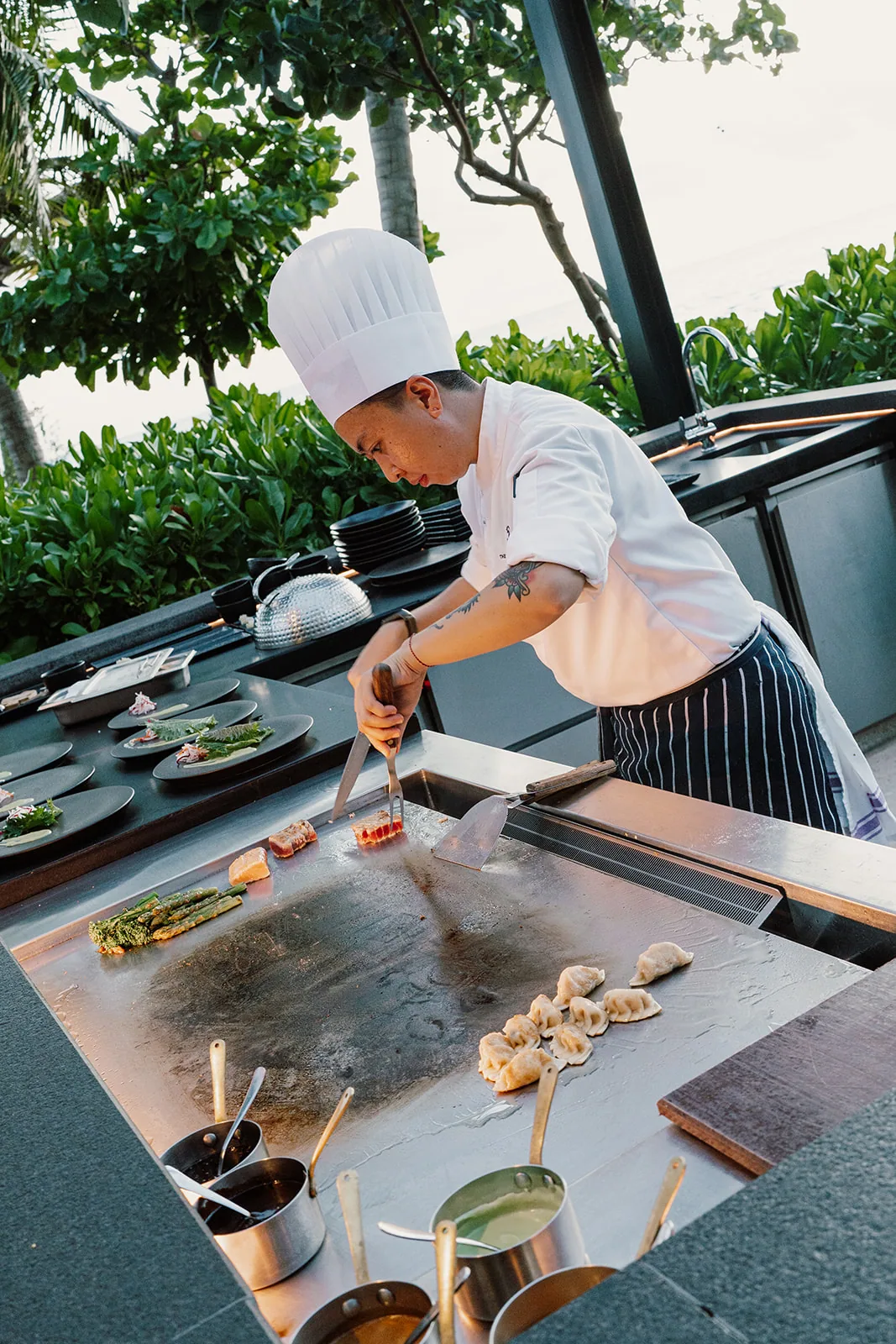 The Ritz-Carlton Maldives, Fari Islands, dining, North Malé Atoll, Maldives, Iwau teppanyaki detail: chef in tall whites plating gyoza dumplings, beef, and grilled greens on the steel grill, plated greens and a stack of dark dining plates set ready, striped chef apron and copper sauce pans below, palm canopy filtered light.