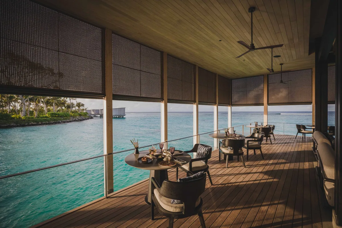 The Ritz-Carlton Maldives, Fari Islands, dining, North Malé Atoll, Maldives, La Locanda over-water dining pavilion: dark wood deck, woven dark-rattan armchairs paired with round breakfast tables, full-height timber-slatted screens framing a wide lagoon view, palm trees and turquoise water visible through the opening, ceiling fans overhead.