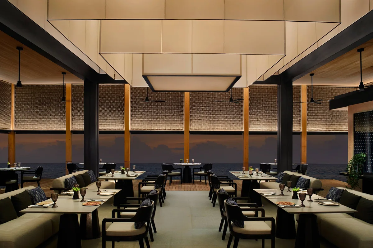The Ritz-Carlton Maldives, Fari Islands, dining, North Malé Atoll, Maldives, Summer Pavilion, the Cantonese dining room: sage-green banquette seating paired with dark dining chairs, dramatic suspended fabric-layered pendant lamps overhead, woven natural-fibre screens lining the walls, full-glass window walls opening onto a dusk-lit ocean, copper goblets on set tables.