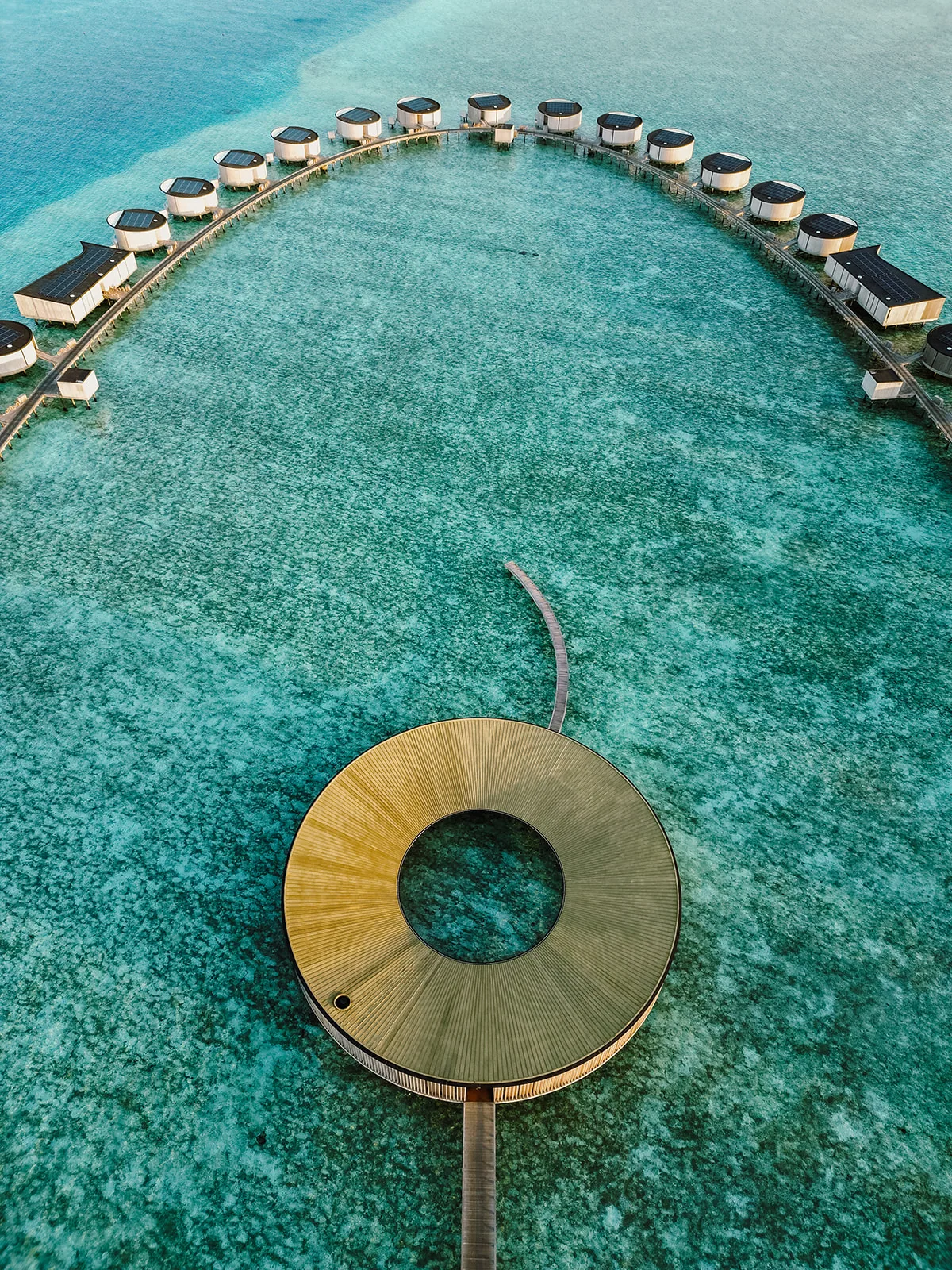 The Ritz-Carlton Maldives, Fari Islands, setting, North Malé Atoll, Maldives, The Ritz-Carlton Maldives Fari Islands wider drone aerial: circular spa pod at the foreground with the half-arc of the over-water villa ring curving across the lagoon, connecting jetty and turquoise reef edge, North Malé Atoll.