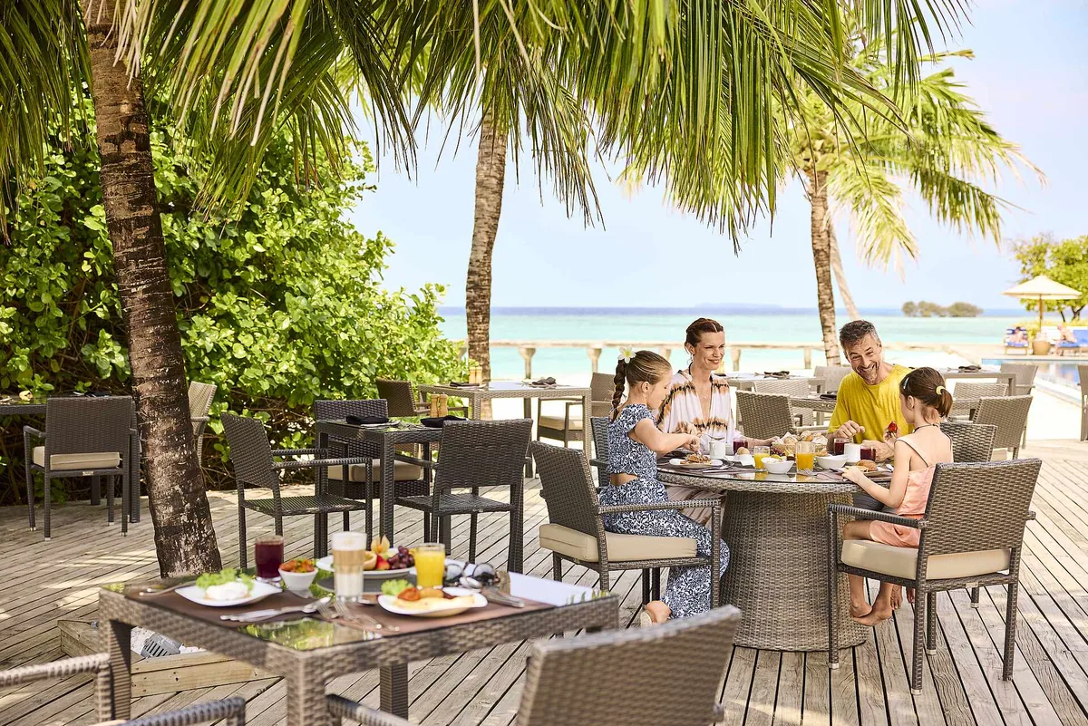 Robinson Club Noonu, main restaurant outdoor breakfast deck under palms with families seated and beach access to the southern sand spit, Noonu Atoll, Maldives