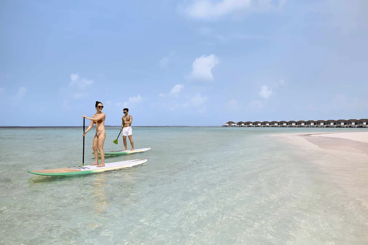 Robinson Club Noonu, couple stand-up paddleboarding on shallow lagoon with overwater villa jetty arm along the eastern reef edge, Noonu Atoll, Maldives