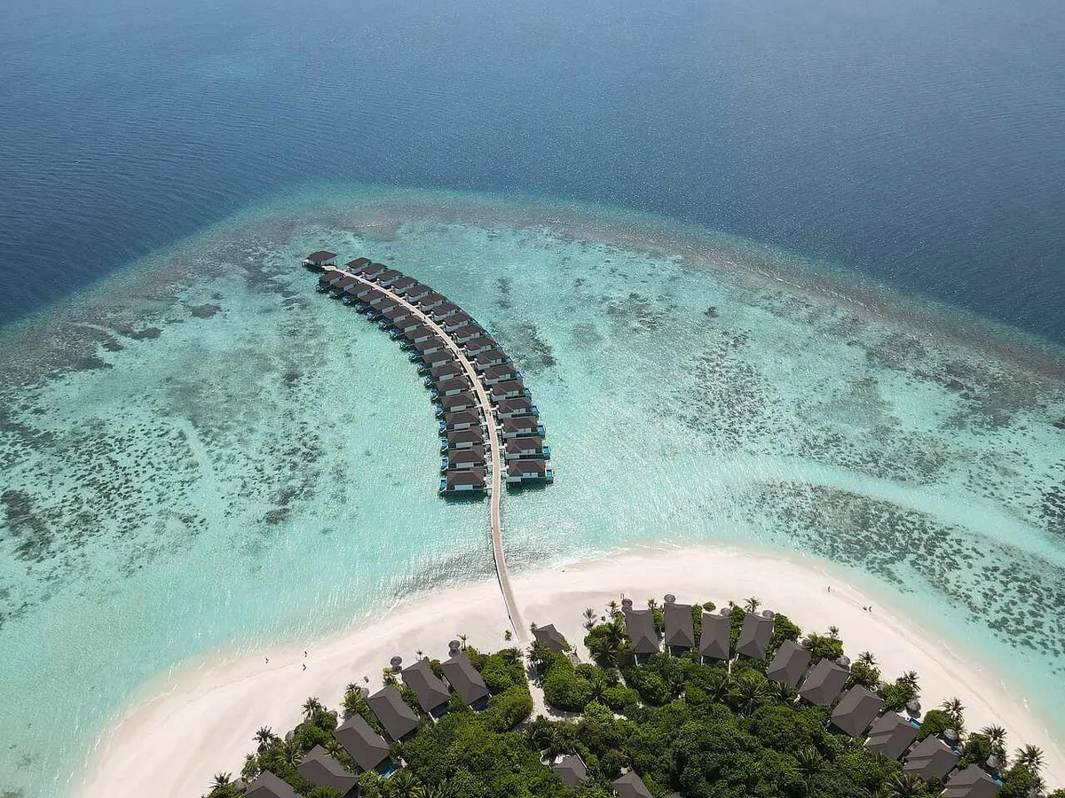 Robinson Club Noonu, aerial of overwater villa jetty arm with adjacent beach villa cluster and southern sand spit on turquoise lagoon, Noonu Atoll, Maldives