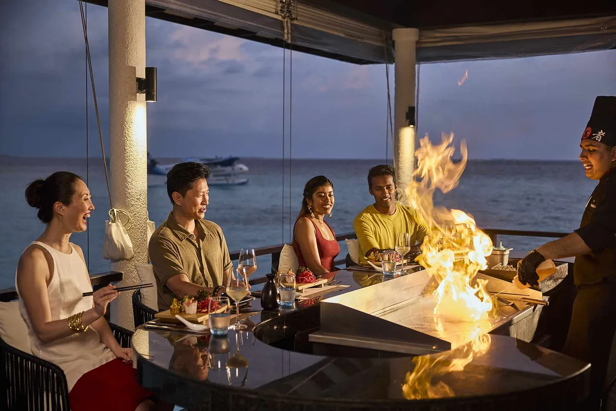 Robinson Club Noonu, Teppanyaki chef flame-cooking at overwater pavilion counter with seaplane moored in the lagoon at dusk, Noonu Atoll, Maldives