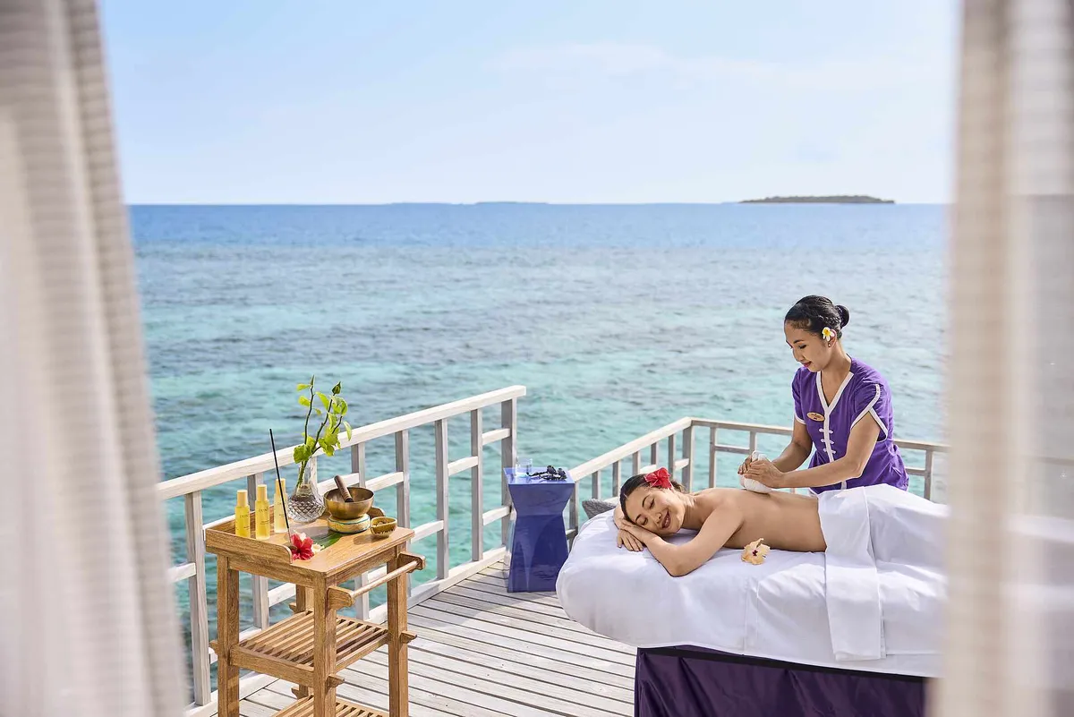Robinson Club Noonu, Duniya Spa therapist applying herbal-poultice massage on overwater treatment deck with distant island on the horizon, Noonu Atoll, Maldives
