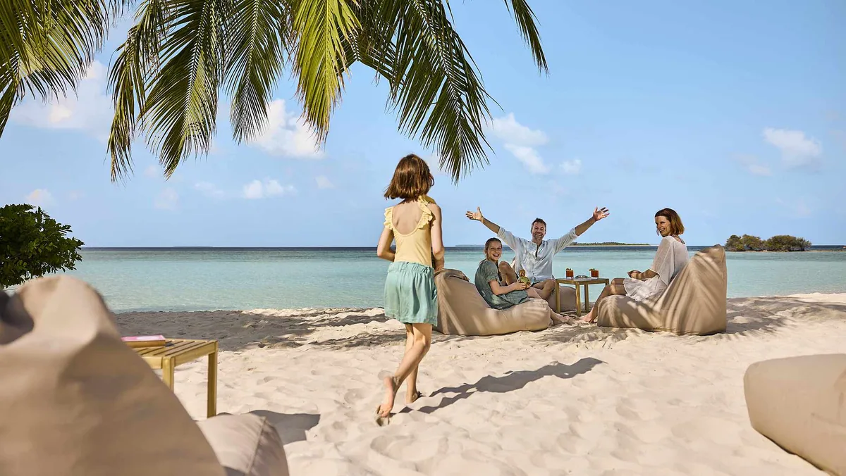 Robinson Club Noonu, Sundowner peninsula beach with bean-bag loungers under coconut palms and adjacent island visible across lagoon, Noonu Atoll, Maldives