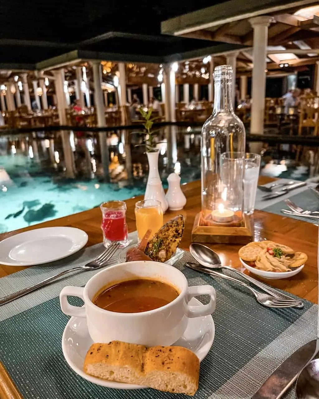Royal Island Resort & Spa, dining, Baa Atoll, Maldives, Maakanaa main restaurant at night, the white-columned colonnaded thatched-roof pavilion reflected on the central reflection pool, table setting in the foreground with bread and soup-bowl on woven placemat, candle lantern and the dining room beyond.