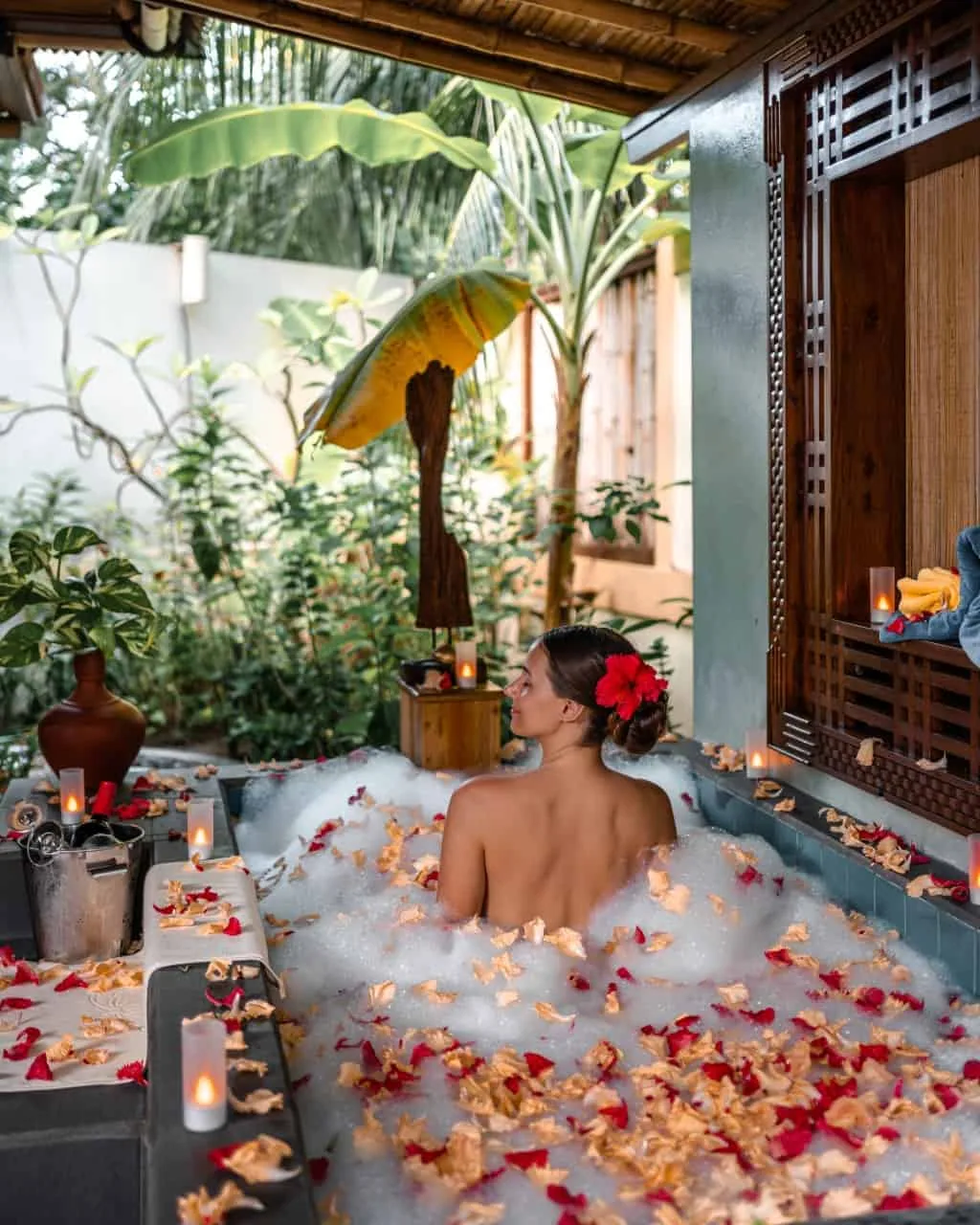 Royal Island Resort & Spa, spa, Baa Atoll, Maldives, Araamu Spa flower-bath setup, the outdoor bathtub strewn with red-and-orange petals and bubble foam beneath the bamboo-eave roof, intricately-carved dark-wood Maldivian screen wall, dense banana-and-tropical-jungle planting, candle altar and silver champagne-bucket setting.