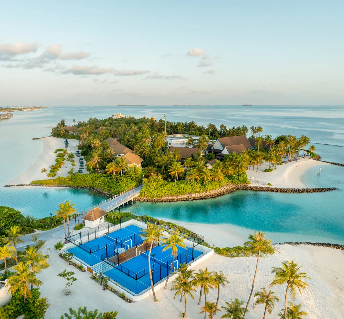 SAii Lagoon Maldives, Curio Collection by Hilton, activity, South Malé Atoll, Maldives, Aerial of the paddle and pickleball sport courts: two blue acrylic courts beside a palm-fringed sand garden, the lagoon and reef edge in the upper frame, the sports-facility programme at the Crossroads cluster shared-services depth.