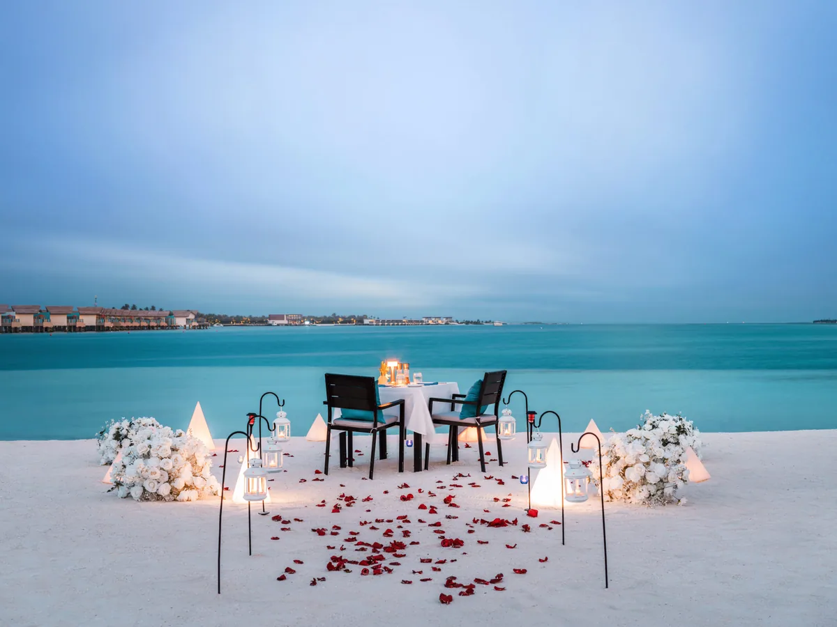 SAii Lagoon Maldives, Curio Collection by Hilton, dining, South Malé Atoll, Maldives, Private beach dinner setup at twilight: table for two on white sand with candle lanterns and conical glass markers, rose-petal path leading up, white floral arrangements flanking the setup, the Crossroads cluster OWVs and Marina precinct buildings visible across the still lagoon under a pale-blue post-sunset sky.