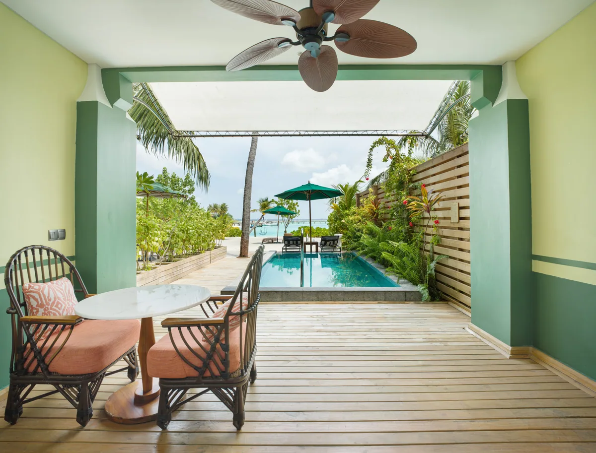 SAii Lagoon Maldives, Curio Collection by Hilton, villa beach, South Malé Atoll, Maldives, Beach Room with Pool: sage-green walls with palm-leaf ceiling fan overhead, terracotta-cushioned wicker chairs at a marble table, sliding doors opening onto a private plunge pool deck with sun-loungers and parasol, lagoon visible beyond.