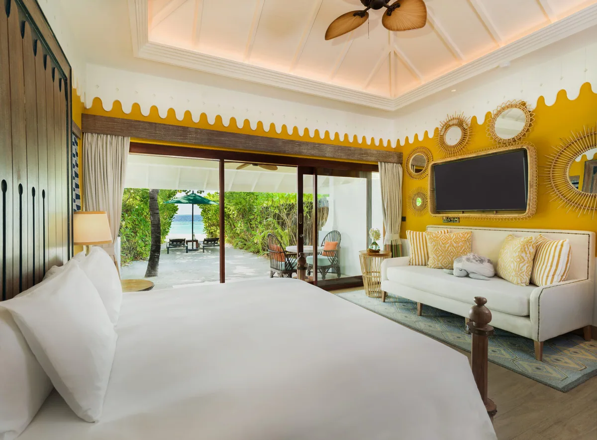 SAii Lagoon Maldives, Curio Collection by Hilton, villa beach, South Malé Atoll, Maldives, Beach Villa King bedroom: signature SAii yellow-scallop accent wall, woven-rattan mirror collection clustered around the wall-mounted TV, white sofa with yellow-pattern accent cushions, white-linen bed, sliding doors opening to the beach with green parasol and twin loungers.