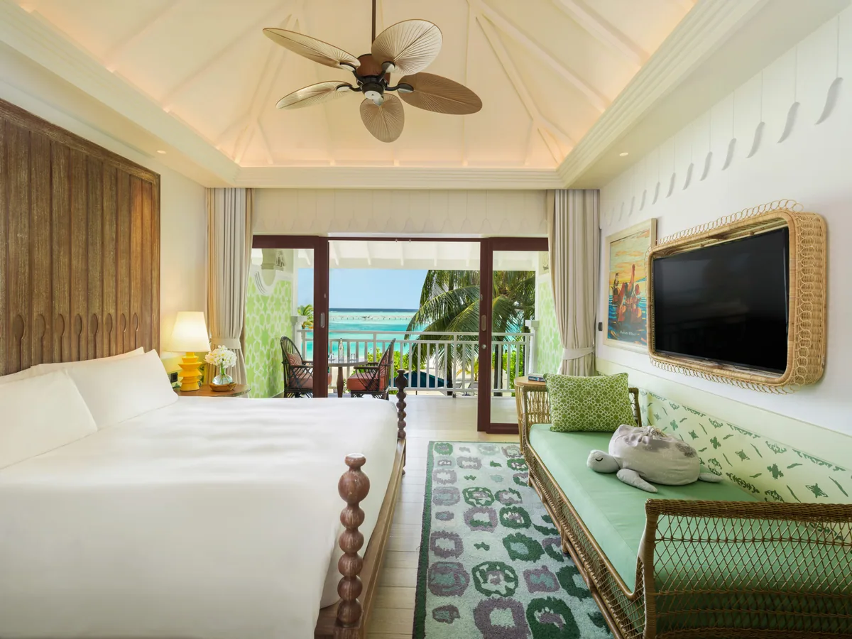 SAii Lagoon Maldives, Curio Collection by Hilton, villa beach, South Malé Atoll, Maldives, Sky Room bedroom at the upstairs tier: white peaked ceiling with palm-leaf fan, woven daybed with green-stencil pillows, light-wood floor, white-linen bed with carved bedposts, French doors opening to a private balcony with green Moroccan-tile screen and lagoon view.