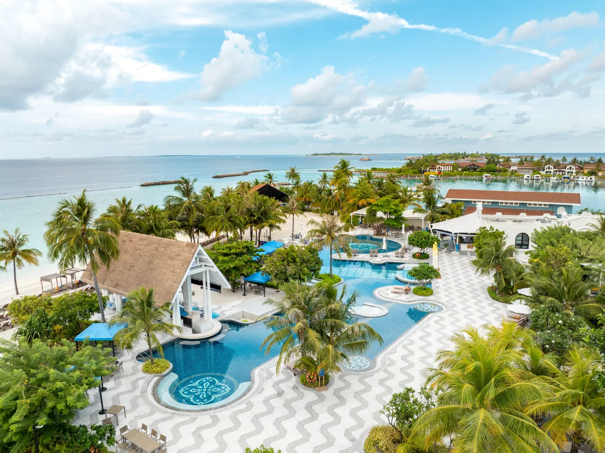 SAii Lagoon Maldives, Curio Collection by Hilton, setting, South Malé Atoll, Maldives, Beach Club aerial: the large connected infinity pools with geometric-pattern paving, palm canopy ringing the pool deck, blue sun-loungers arrayed at the pool edge, the Marina precinct buildings (white with red-tile roofs) visible across the lagoon at the cluster centre.
