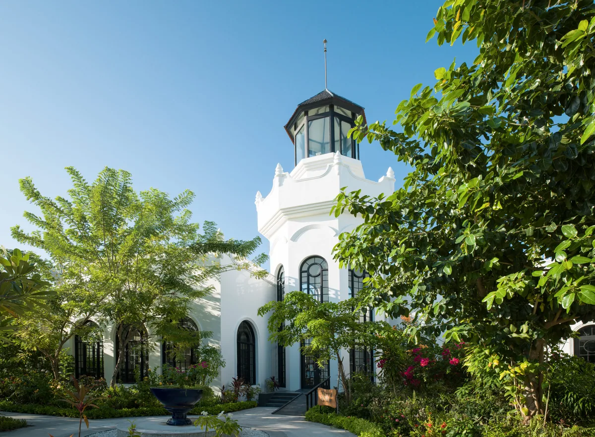 SAii Lagoon Maldives, Curio Collection by Hilton, setting, South Malé Atoll, Maldives, Beach Club exterior architecture: white colonial-revival pavilion with a slim glass cupola tower, arched windows, lush tropical garden framing the entrance, the distinctive landmark of the SAii Lagoon arrival experience.