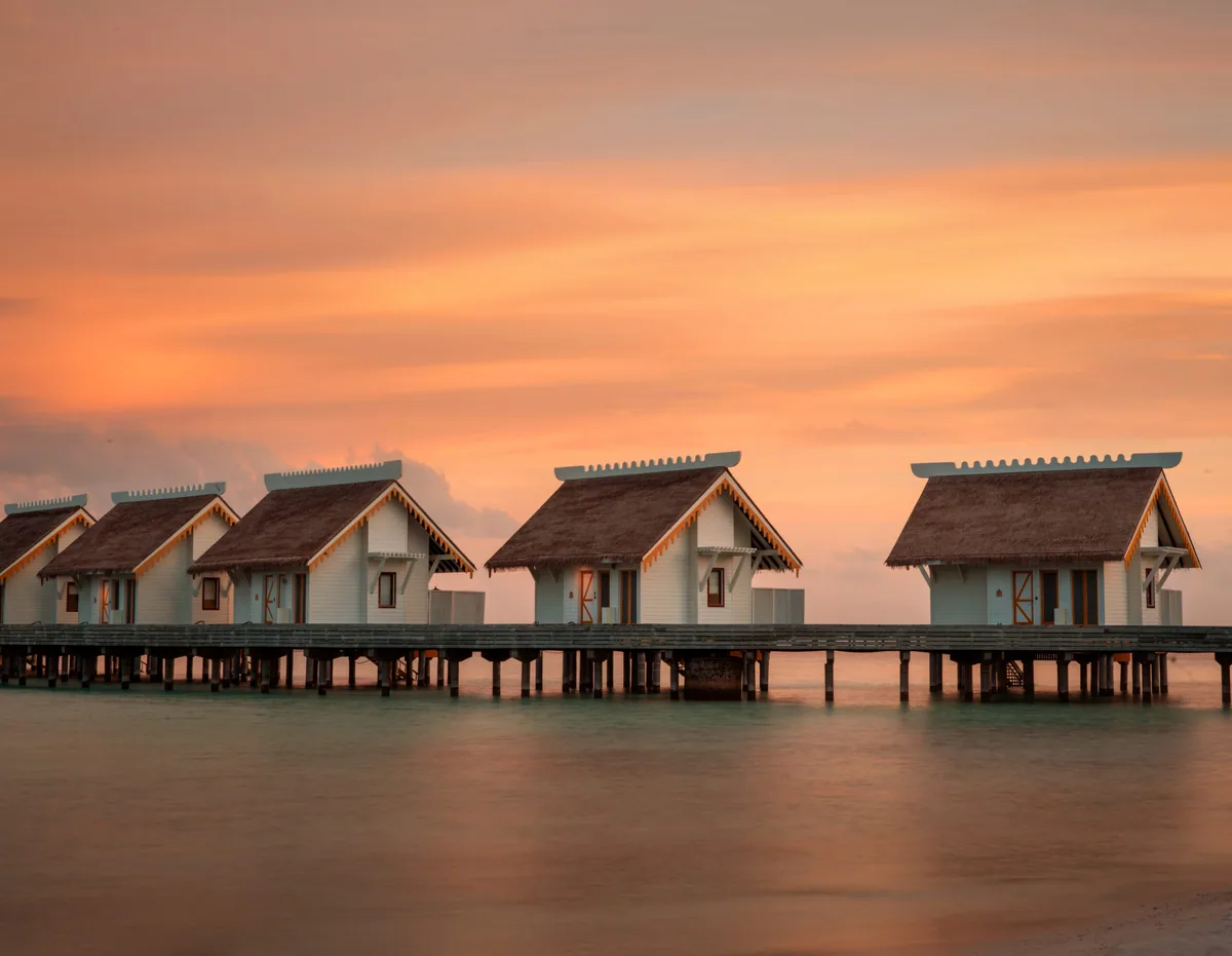 SAii Lagoon Maldives, Curio Collection by Hilton, villa overwater, South Malé Atoll, Maldives, Overwater villa row at sunset: line of peaked thatched-roof villas with the signature yellow trim on stilts above the calm sunset-mirrored lagoon, dramatic peach and orange sky behind, the OWV inventory at its golden-hour identity.