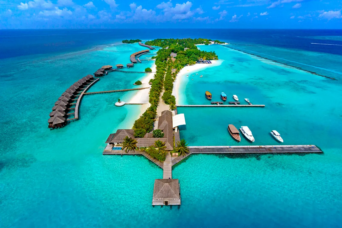 Sheraton Maldives Full Moon Resort & Spa, aerial, North Malé Atoll, Maldives, Full-island approach aerial: Furanafushi seen from the channel side with the T-shaped arrival jetty and pavilion at the foreground, wooden dhonis and Sheraton speedboats moored, central island palm canopy, full curve of the namesake thatched-roof overwater villa row tracing the lagoon edge into the distance.