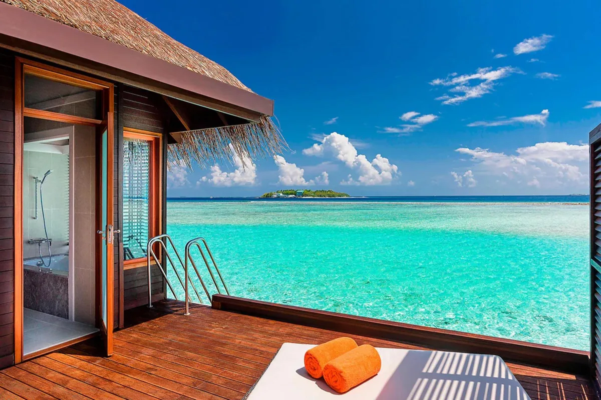 Sheraton Maldives Full Moon Resort & Spa, villa overwater, North Malé Atoll, Maldives, Water Bungalow deck POV: thatched-roof corner overhead, glass door and picture window framing the in-villa bathroom on the left, stainless ladder descending from the deck into the turquoise lagoon, two rolled orange-towel loungers on the timber deck, small distant green island on the horizon (the property's signature thatched-roof and step-down-into-lagoon configuration).