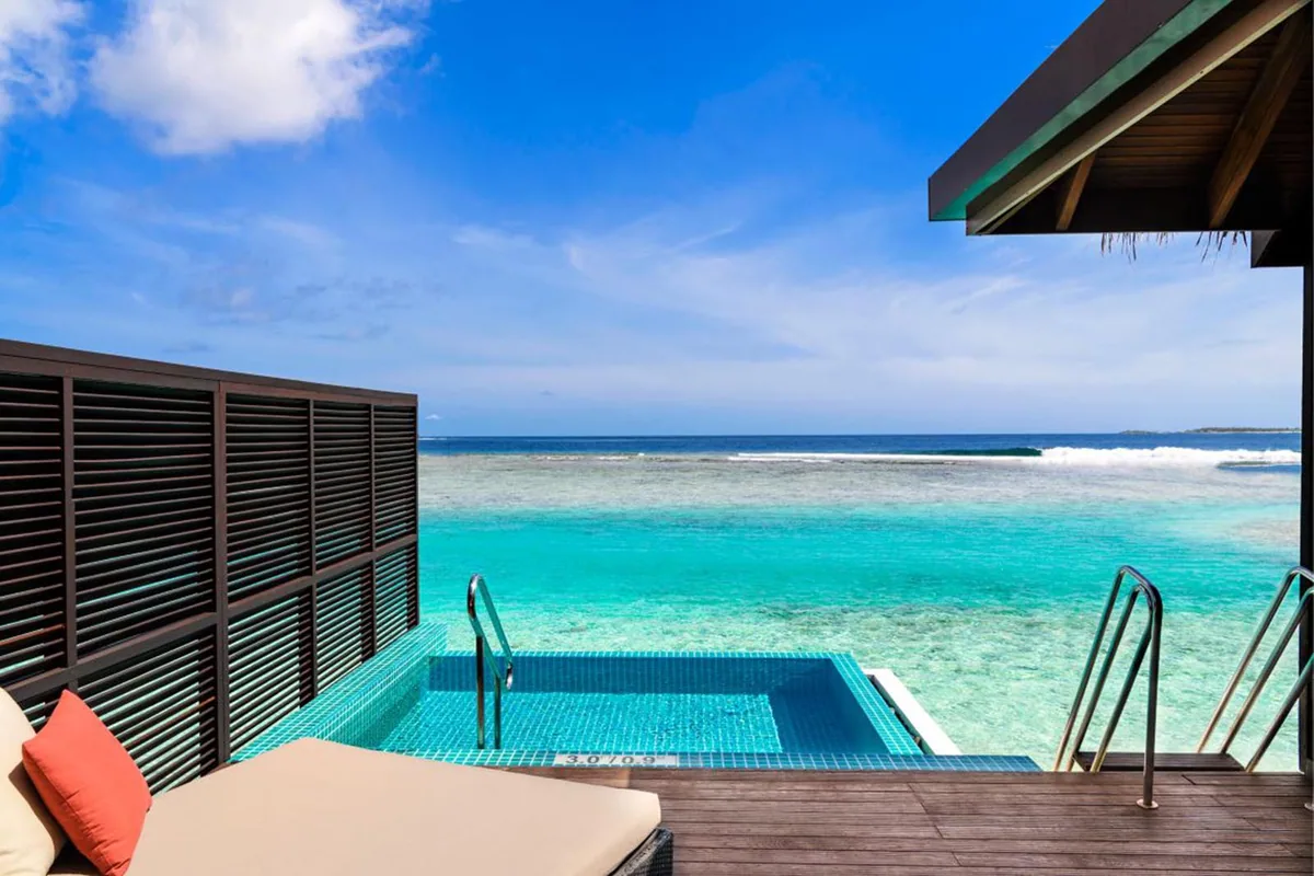Sheraton Maldives Full Moon Resort & Spa, villa overwater, North Malé Atoll, Maldives, Overwater Villa with Pool deck: dark-louvered villa wall on the left, blue-tile plunge pool with submerged ladder built into the timber deck, orange-cushion lounger in the foreground, reef edge and breakwater visible at the horizon, partial thatched roof corner upper-right.