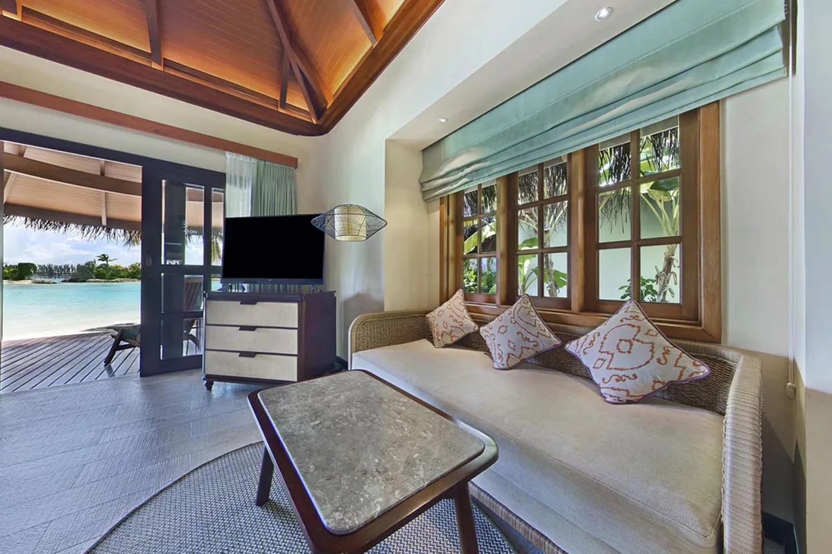 Sheraton Maldives Full Moon Resort & Spa, villa beach, North Malé Atoll, Maldives, Beach Front Cottage interior: vaulted dark-wood ceiling with central beam structure, daybed banquette under multi-pane wood-framed windows with green Roman shades, batik-print pillows in coral-and-cream, woven-front credenza with wall-mounted television, sliding glass door at left opening to the deck with beach and lagoon beyond.