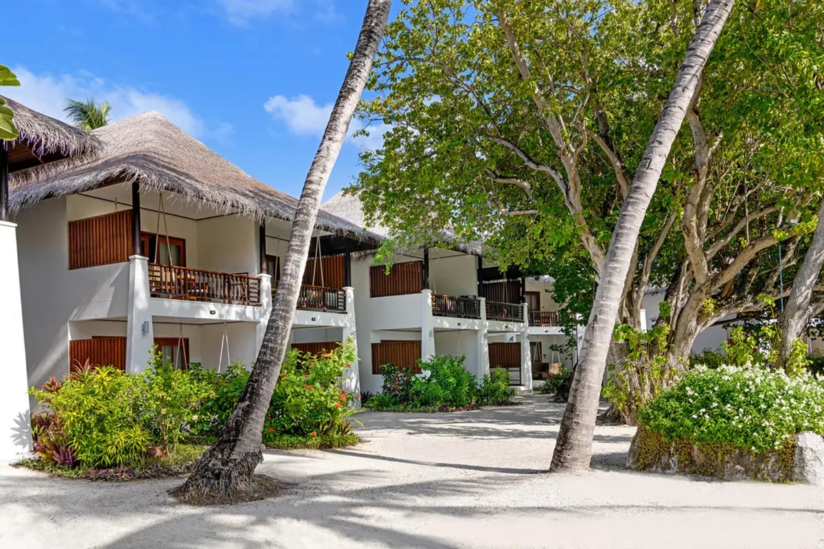 Sheraton Maldives Full Moon Resort & Spa, villa beach, North Malé Atoll, Maldives, Deluxe Room building exterior: two-story accommodation block with thatched canopy roof feature over the entry corridor, balconied upper-floor rooms, palm trees framing the approach, dense planted shrubs at the ground level (architecturally distinct from the heritage single-story thatched bungalows).
