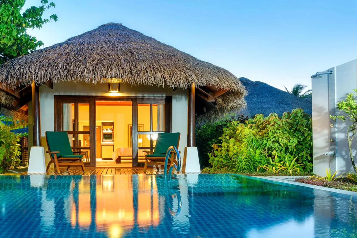 Sheraton Maldives Full Moon Resort & Spa, villa beach, North Malé Atoll, Maldives, Cottage with Pool at twilight: standalone thatched-roof cottage with lit interior visible through open sliding glass doors, two green-cushioned lounge chairs on the timber deck, blue-tile plunge pool in the foreground, planted shrub border at right with outdoor rain shower, secondary cottage thatched roof in the background.