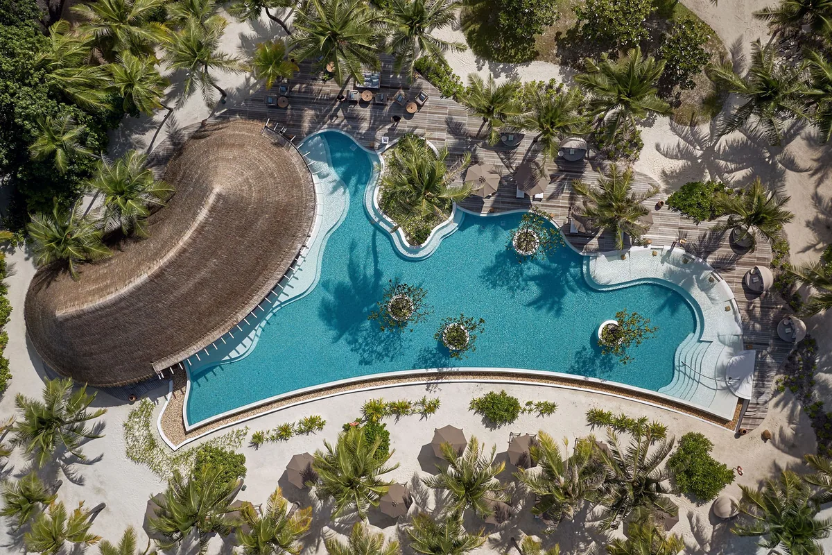 Six Senses Kanuhura, pool, Lhaviyani Atoll, Maldives, Sip and Sand pool aerial top-down: free-form amoeba-shaped main pool with three circular planter-island features within the water, thatched circular roof of the swim-up bar on the left edge, white sand fringe and dense coconut-palm canopy enclosing the pool deck, daybed loungers visible at the periphery.