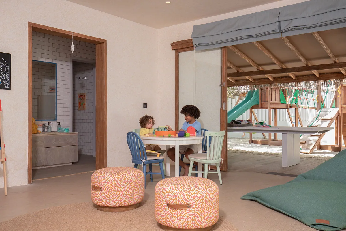 Six Senses Kanuhura, activities, Lhaviyani Atoll, Maldives, Grow With Six Senses Kids Club interior: children at a low round play table with colour-block toys, blue and mint-green Windsor children's chairs, pink-and-orange-patterned floor poufs, washroom with white subway-tile wall visible through the open doorway at left, an outdoor timber climbing-frame-and-slide structure visible through the open glass doors.