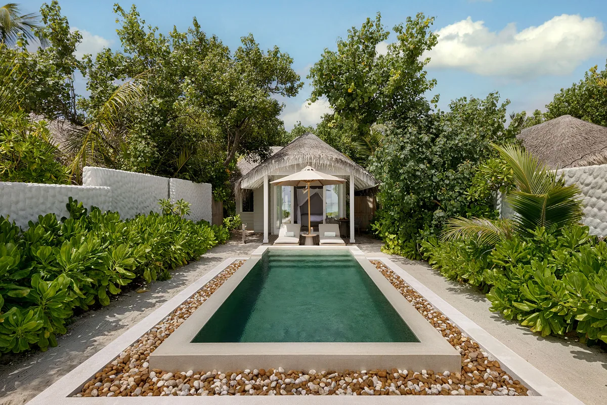 Six Senses Kanuhura, villa beach, Lhaviyani Atoll, Maldives, Beach Pool Villa courtyard: thatched-roof bedroom pavilion at the rear, rectangular plunge pool framed by white pebble-stone decking, dense scaevola hedge walls on both flanks, tropical greenery enclosing the courtyard from the resort circulation.