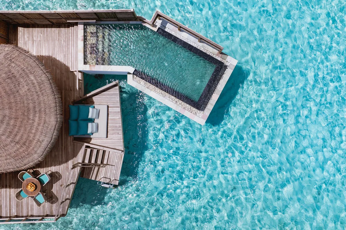 Six Senses Kanuhura, villa overwater, Lhaviyani Atoll, Maldives, Water Villa with Pool top-down aerial: circular thatched roof structure, rectangular overwater plunge pool extending into the lagoon, twin sun loungers on the deck, dining table at the deck edge, turquoise lagoon water visible through the glass-floor cutout.