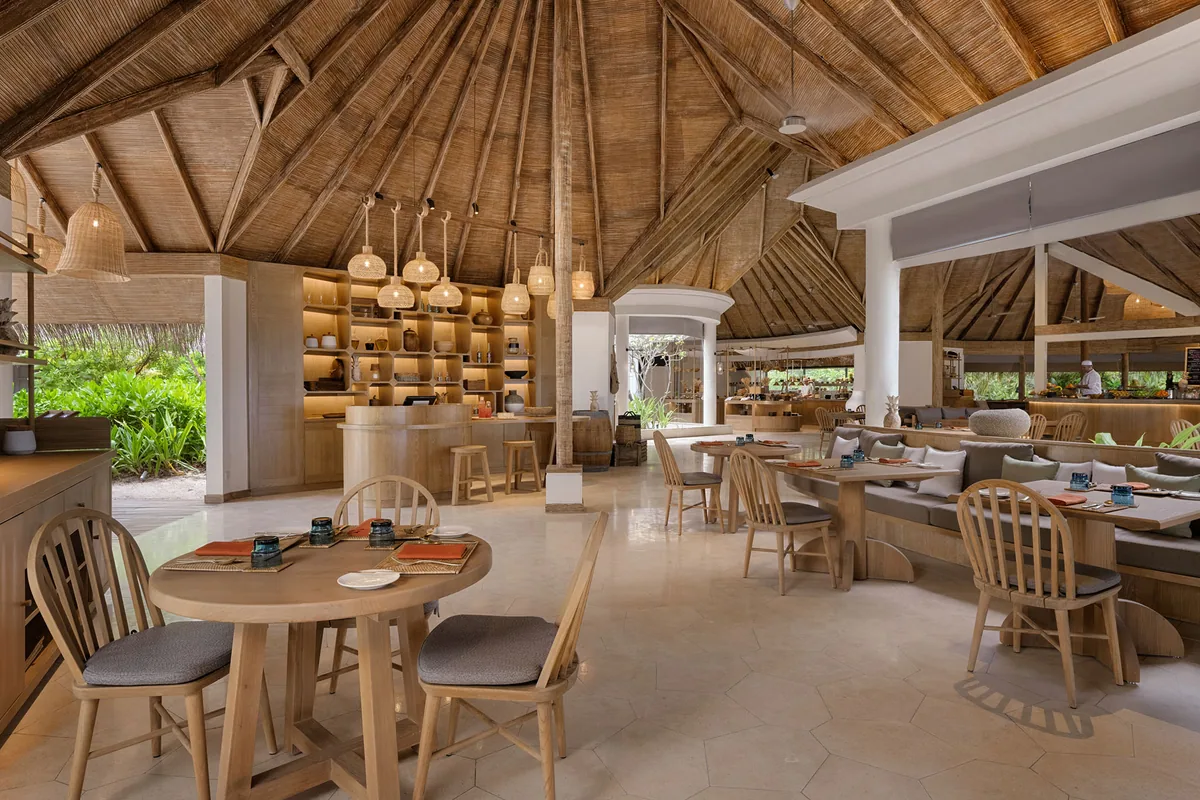 Six Senses Kanuhura, dining, Lhaviyani Atoll, Maldives, The Market principal all-day pavilion: high thatched cathedral roof with exposed radial timber rafters, woven rattan pendant lights, blonde-wood Windsor-style chairs at round tables, central wood-clad open-kitchen counter, banquette seating along the side wall, hexagonal beige floor tile.