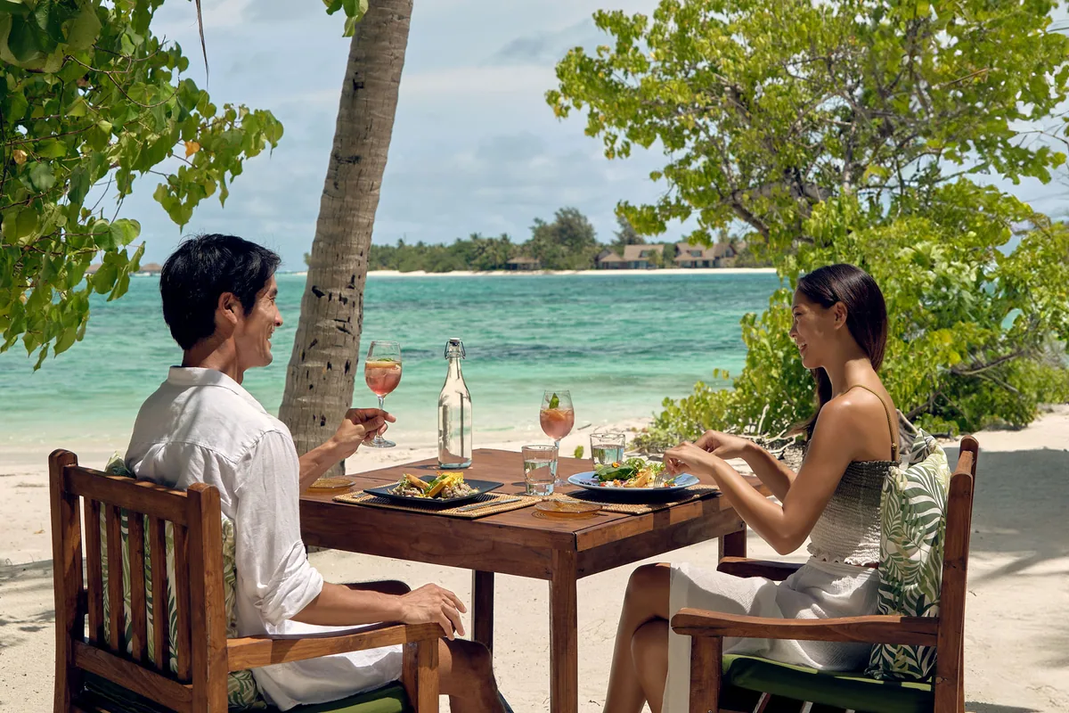 Six Senses Kanuhura, dining, Lhaviyani Atoll, Maldives, Drift castaway beach-dining on Jehunuhura: small wooden table set with linen runners and tropical garnish, feet-in-the-sand position under a coconut-palm canopy, the resort's main island and turquoise channel visible across the lagoon, the Drift signature lunch-and-sundowner programme on the seven-minute-tender-distance private island.