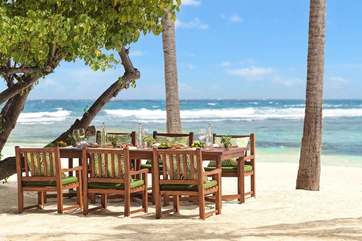 Six Senses Kanuhura, dining, Lhaviyani Atoll, Maldives, Drift Jehunuhura private-island long-table lunch staging: heavy timber communal table for eight with green-striped cushion seating, glassware and place settings laid for service, palm trunks framing the composition, reef-break surf visible across white sand, the castaway-style group-lunch configuration distinct from the small-table beachside service.