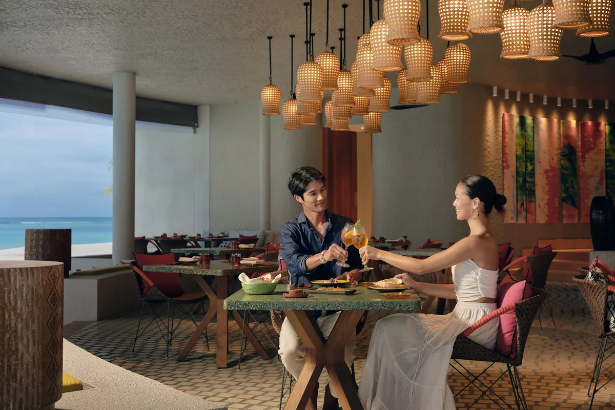 Six Senses Kanuhura, dining, Lhaviyani Atoll, Maldives, The Point Spanish-tapas-and-pintxos restaurant: open-air pavilion under a concrete-slab roof, signature cluster of dozens of woven-cane basket-form pendant lights suspended at varied heights, jade-green terrazzo tabletops with cross-leg timber bases, woven rattan chairs with red and pink cushions, ocean view through the columns to the left.