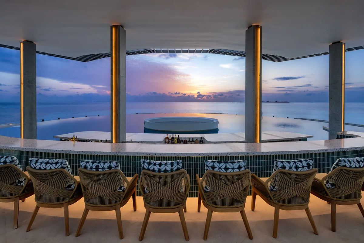 Six Senses Kanuhura, bar, Lhaviyani Atoll, Maldives, Sunset Point rooftop bar at twilight: open-air pavilion framed by concrete columns with warm uplighting, curved terrazzo bar counter, woven-rope bar stools cushioned with blue-and-white batik pillows, a sunken circular jacuzzi feature in the foreground infinity pool, peach-and-lavender sunset sky over the horizon ocean.